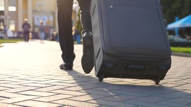 Feet of successful businessman walking through city street and pulling suitcase on wheels at sunny day. Confident man on way to his flight. Male legs in shoes stepping at urban environment. Slow mo - Footage, Video