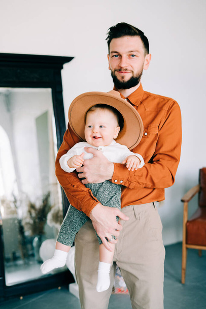 um jovem pai elegante em tendência camisa laranja brinca com seu filho em um chapéu marrom em seus braços em um estúdio elegante espaçoso
. - Foto, Imagem
