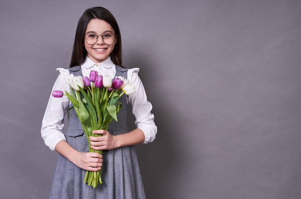 Graceful cute emotional lady long-haired brunette in glasses, holding tulips in the hands of many-colored multi-colored happy gift to the left of copy space, very joyful, on a gray background. - Foto, imagen