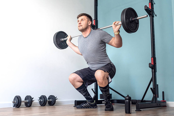 Sports Man Doing Squats With Barbell Row at Home in His small and Bright Apartment with Minimalistic Interior. Quarantine. - Фото, изображение
