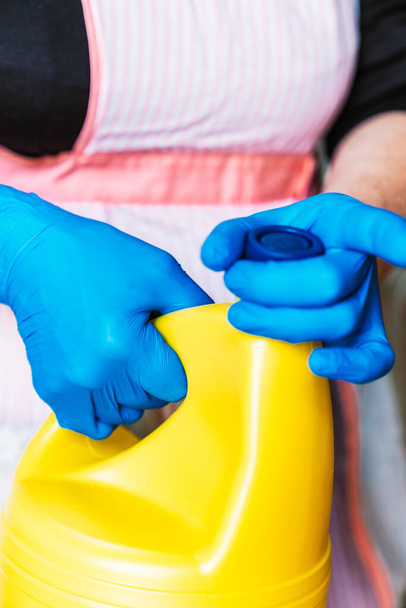 Woman with blue latex gloves and apron opening a yellow bottle of bleach. - Foto, Imagem