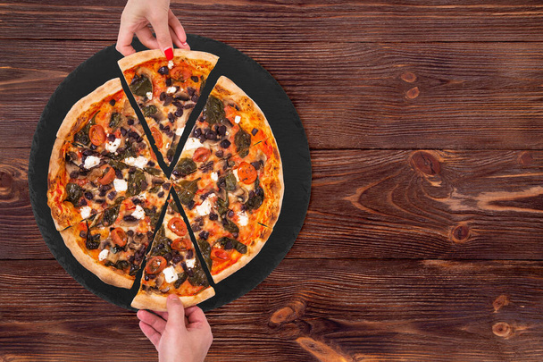 Junge Leute nehmen leckere Pizzascheiben mit Kirschtomaten, Spinat, Mozzarella, Feta, Kalamata-Oliven und Pilzen von der schieferschwarzen Platte, die auf einem rustikalen Holztisch steht, Platz für Text. - Foto, Bild
