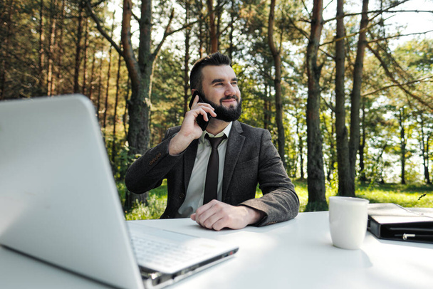 Joven hombre de negocios atractivo en traje y corbata se sienta en el escritorio y trabaja en la computadora al aire libre. Bebe café de taza blanca y habla por teléfono. Árboles verdes, naturaleza y parque en el fondo
. - Foto, Imagen