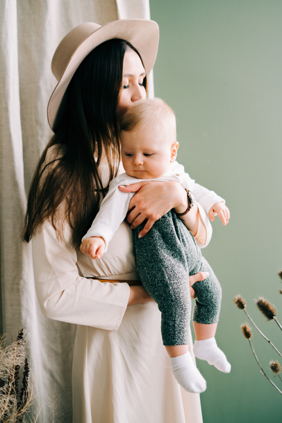 jonge moeder in trend beige jurk en hoed, met haar stijlvol geklede zoon in haar armen in een ruime Studio. - Foto, afbeelding