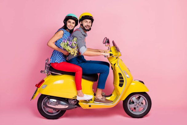 Full body profile side photo of positive motorcyclists two people drivers riders drive yellow fast scooter enjoy rural field adventure hold wildflowers isolated over pink color background - Фото, зображення