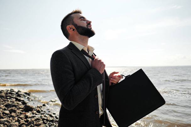 Young attractive male businessman in a brown suit with a briefcase stands on the seashore. Unfastens shirt, loosens tie and raises hands to sea. - Valokuva, kuva