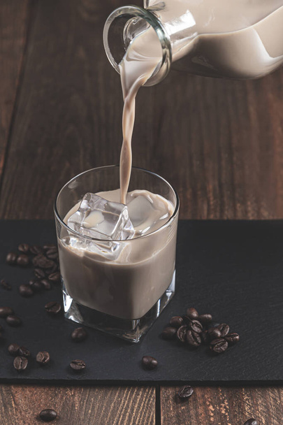Pouring  Cream coffee liqueur in a glass with ice, surrounded by coffee beans on a dark wooden background. - Photo, image