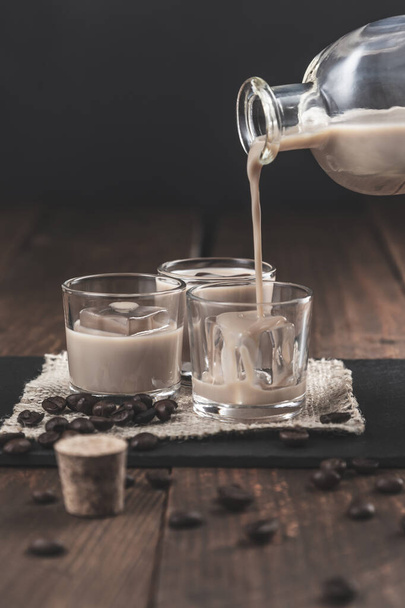 Despejando licor de café Creme em copos com gelo, cercado por grãos de café em um fundo de madeira escura
. - Foto, Imagem