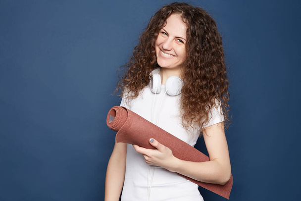 Portrait of attractive Caucasian curly haired woman  dressed in sports clothing, holding brown yoga mat,wearing headphones, listens to music, poses on ultramarine studio wall, copy space area around. - Photo, Image