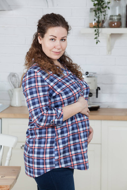 Portrait of pregnant woman holding her belly, dressed plaid shirt, curly long hair. Standing in kitchen interior - Valokuva, kuva