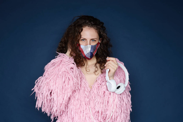 Home party concept. Attractive curly haired female ,wearing protective colorful fashionable mask, fluffy jacket, holding headphones, looking at camera with blue eyes, isolated indoors on ultramarine. - Photo, Image