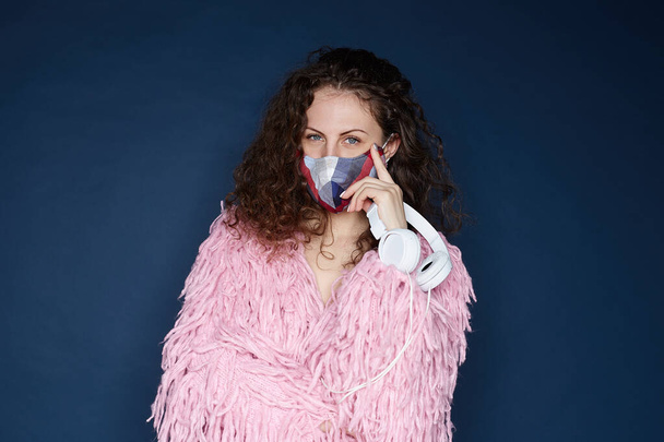 Home party concept. Attractive curly haired female ,wearing protective colorful fashionable mask, fluffy jacket, holding headphones, looking at camera with blue eyes, isolated indoors on ultramarine. - Photo, Image