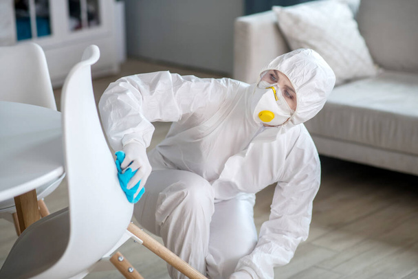 Woman in white workwear and gloves disinfecting the chair - Фото, изображение
