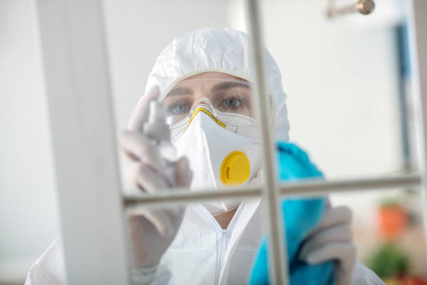 Woman in white workwear and respirator disinfecting the window - Фото, изображение