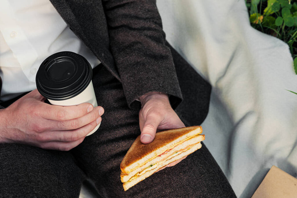 Manos de un hombre en traje con un vaso de café para llevar y un sándwich triangular con atún
. - Foto, Imagen