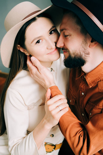 un couple amoureux dans des vêtements branchés et des chapeaux sont assis sur un canapé marron, embrassant dans un beau Studio spacieux
. - Photo, image