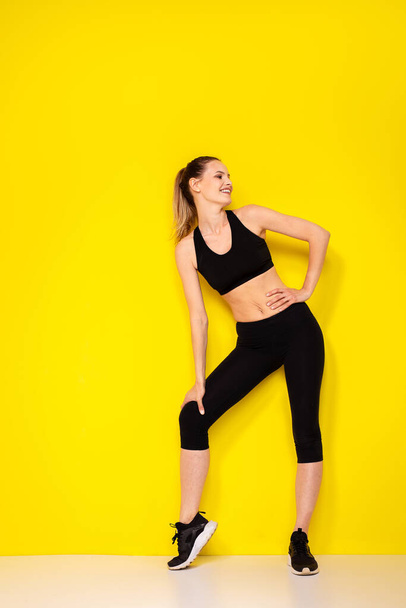young and athletic woman in sportwear smiling and being happy of her body and her lifestyle on yellow background - Photo, Image