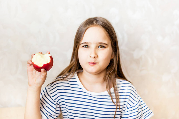 A girl with long hair in a striped T-shirt sits at a desk at home and eats between online lessons, learning via the Internet, wholesome food, body growth, vitamins, beautiful teeth - Valokuva, kuva