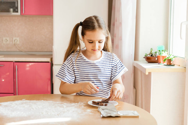 A girl with long hair in a striped t-shirt in the kitchen prepares chocolate for baking, flour, plates and a rolling pin for baking, croissants - Photo, image