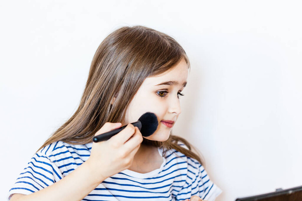 a little girl sits at a table and paints her eyes and lips with her mother's makeup, while her mother is not at home, for the first time trying makeup, creating beauty on her face, coronavirus - 写真・画像
