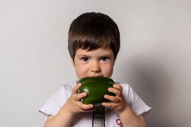 Een jongetje houdt een groene peper in zijn handen en kijkt goed. Groene groenten in het dieet van kinderen, het gebruik van goede voeding voor kinderen. - Foto, afbeelding