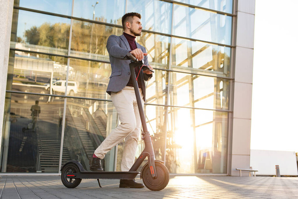 Young handsome stylish man with e-scooter standing on sidewalk near the business center on the sunset. Trendy urban transportation on modern electric scooter. Eco friendly mobility concept - Фото, изображение