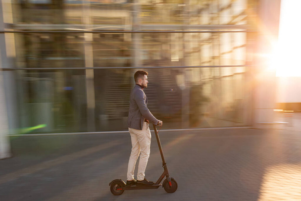 Young handsome man riding the e-scooter on the road near the airport on the sunset. Trendy urban transportation on modern electric scooter. Eco friendly mobility concept - Foto, Bild