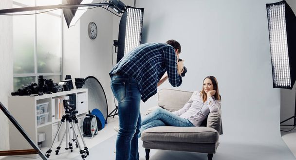 Professional photo shoot at the studio, a female model is posing on an armchair and smiling; the photographer is taking pictures with his digital camera - Фото, зображення