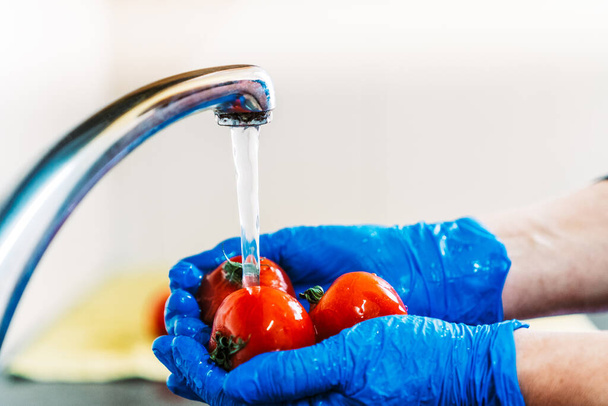 Hands with blue latex gloves disinfecting tomatoes to decontaminate the fruit from coronavirus. Washing the fruit with water in the kitchen sink with the tap. - Foto, Imagem