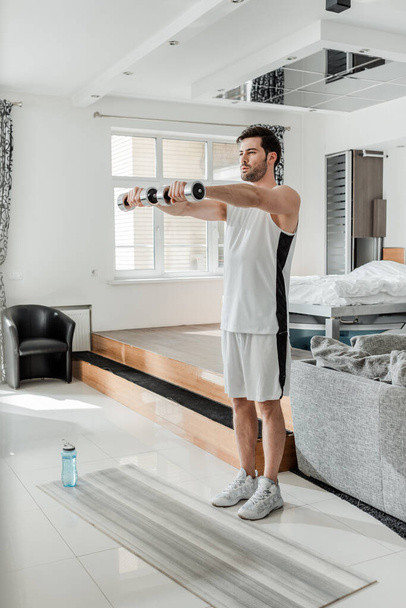 Man in sportswear exercising with barbells near sports bottle and fitness mat at home  - Photo, Image