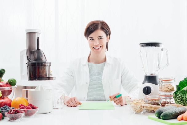Smiling professional nutritionist holding an apple, she has healthy fruits, vegetables and juicers on her desk; healthy diet and wellness concept - Foto, Imagem
