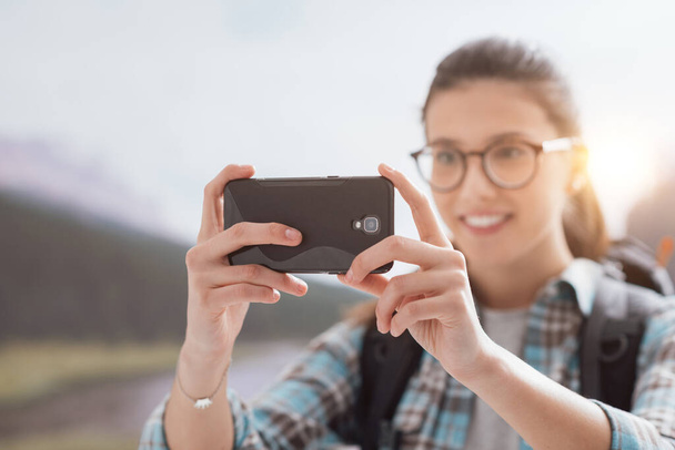 Jovem mulher sorridente caminhando na natureza nas montanhas e tirando fotos com seu smartphone touch screen
 - Foto, Imagem