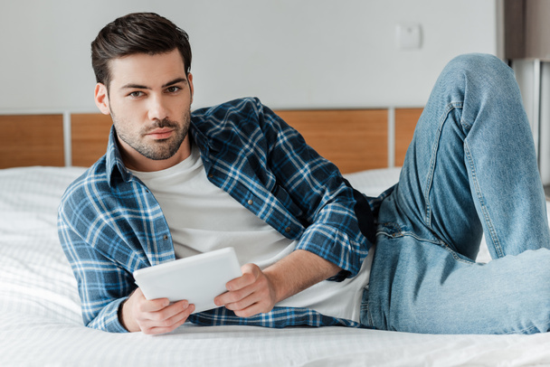 Handsome man in plaited shirt and jeans holding digital tablet ans looking at camera on bed  - Foto, imagen