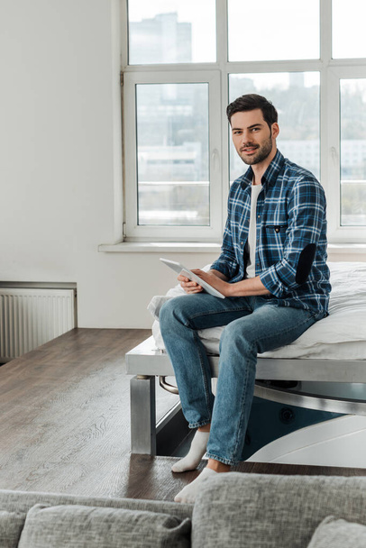 Selective focus of handsome man in checkered shirt looking at camera while holding digital tablet on bed  - Fotografie, Obrázek
