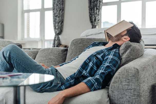 Selective focus of man lying on couch with book on face at home  - Фото, зображення