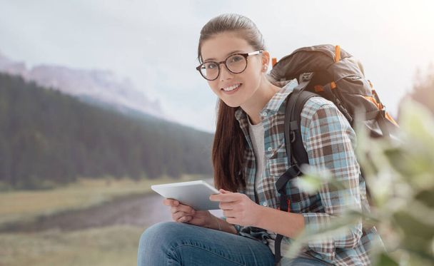 Smiling young girl hiking on the mountains, she is relaxing and connecting with a digital tablet - Foto, Bild