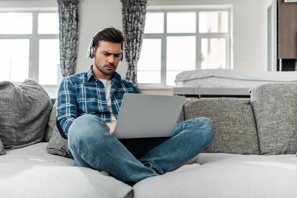 Guapo freelancer usando laptop y escuchando música en auriculares en sofá
  - Foto, imagen