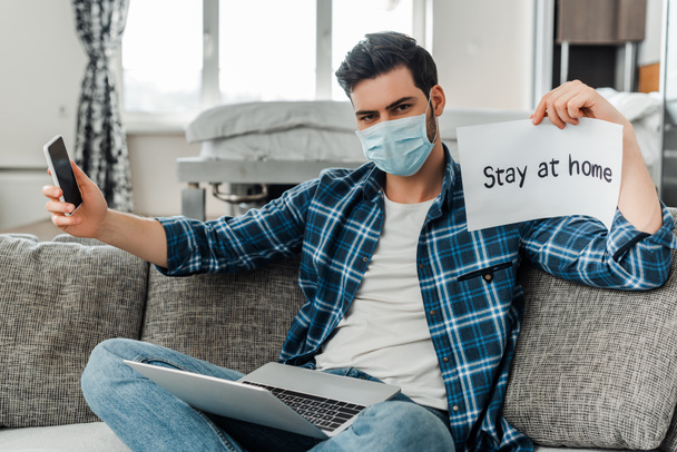 Man in medical mask holding smartphone, card with stay at home lettering and laptop on couch  - Foto, Bild