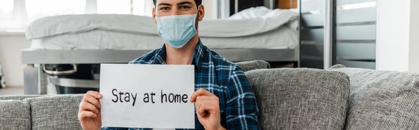 Panoramic orientation of man in medical mask holding card with stay at home lettering on sofa at home  - Фото, зображення