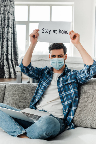 Freelancer in medial mask holding card with stay at home lettering near laptop on couch  - Фото, зображення