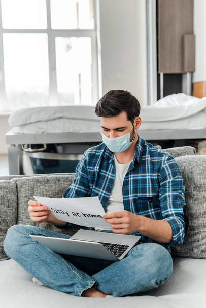 Freelancer in medical mask holding card with stay at home lettering and laptop on sofa  - Foto, immagini