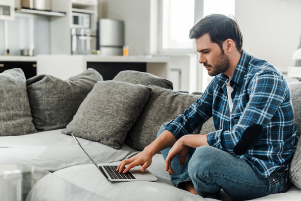 Oldalnézet jóképű férfi használja laptop kanapén otthon  - Fotó, kép