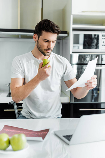 Selective focus of handsome man working with papers and holding apple near laptop in kitchen  - Zdjęcie, obraz