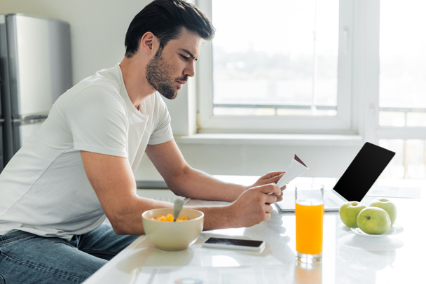 Side view of handsome man holding paper near cereals and orange juice on kitchen table  - Фото, изображение
