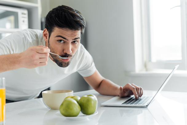 Enfoque selectivo del hombre mirando a la cámara mientras desayuna y usa el portátil en la cocina
  - Foto, Imagen