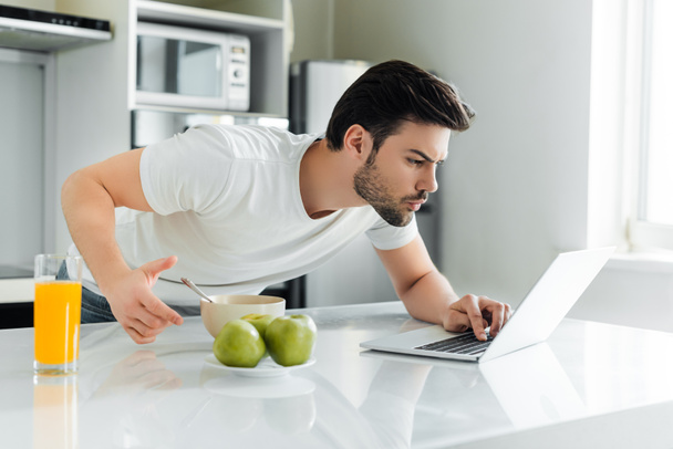 Selective focus of man using laptop near orange juice, apples and bowl on kitchen table  - Fotoğraf, Görsel