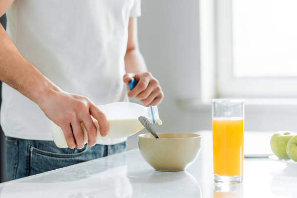 Vista recortada del hombre vertiendo leche en un tazón cerca del jugo de naranja y manzanas en la mesa
  - Foto, Imagen