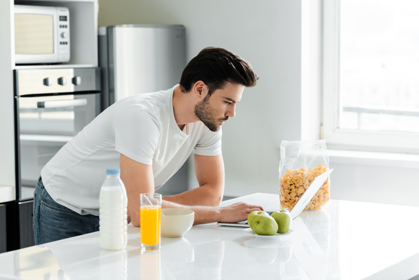 Чоловік використовує ноутбук біля зернових, молока та апельсинового соку на кухонному столі
  - Фото, зображення