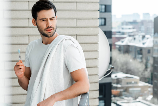 Selective focus of handsome man with toothbrush and towel looking away on balcony at home  - Foto, Bild