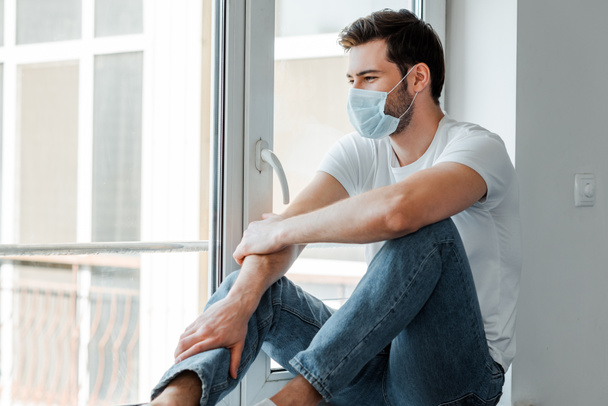 Man met medisch masker zittend op vensterbank thuis  - Foto, afbeelding